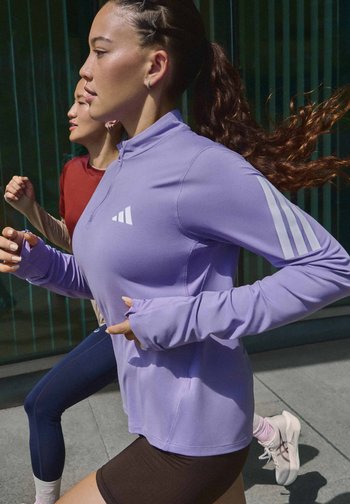 Two women running outdoors wearing athletic gear, one in a purple long-sleeve shirt and black shorts, the other in a red top and navy leggings.