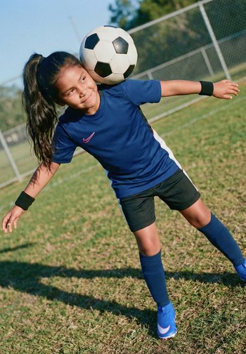Mädchen in blauem Fußballtrikot köpft einen schwarz-weißen Fußball auf einem Grasfeld vor einem Maschendrahtzaun im Hintergrund.