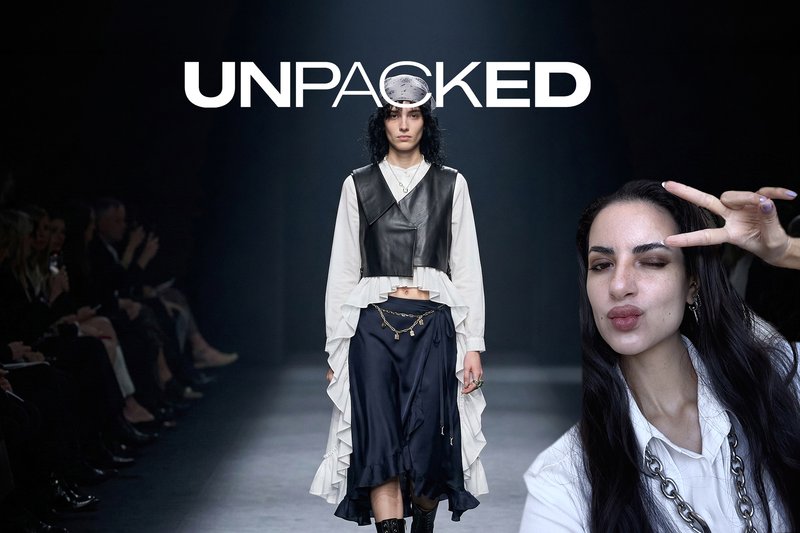 Model walks runway wearing layered white blouse and black leather vest with navy skirt as person poses with peace sign nearby.