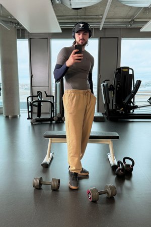 A man in a gym wears a fitted top, casual pants, and headphones, posing with a phone. Dumbbells and kettlebells are on the floor nearby.