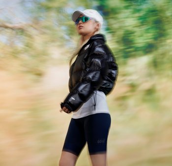 Eine Frau mit langen blonden Haaren, die eine schwarze Daunenjacke, ein weißes Hemd und Sonnenbrillen trägt, geht selbstbewusst vor einem verschwommenen natürlichen Hintergrund.