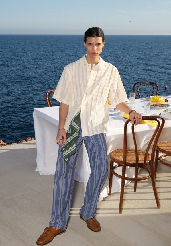 Jeune personne debout près d'une table dressée pour un repas en plein air au bord de la mer, portant une chemise à manches courtes, un pantalon rayé et des chaussures marron.