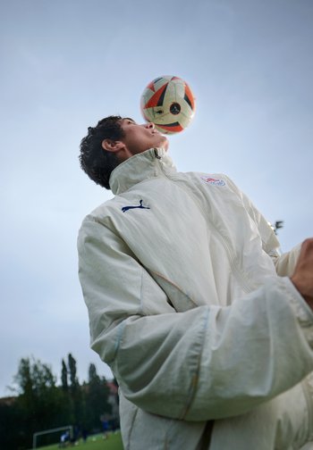 Veste de sport blanche avec col haut ; joueur équilibrant un ballon de soccer sur son front. Champ de gazon vert et ciel nuageux en arrière-plan.