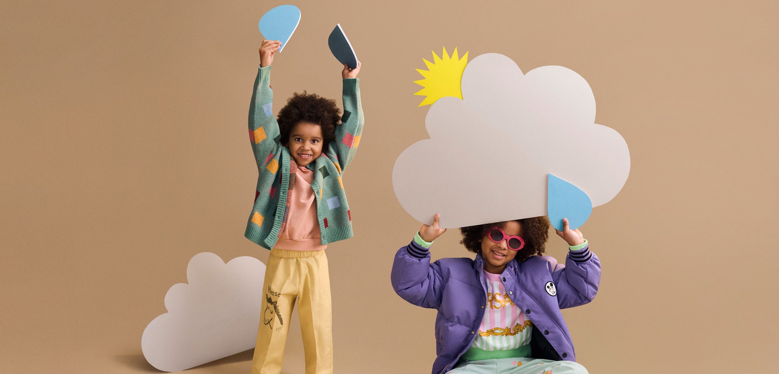 Niño con chaqueta morada y gafas de sol rosas sostiene un recorte de nube con sol y gota de lluvia, sentado con las piernas cruzadas en un suelo beige.