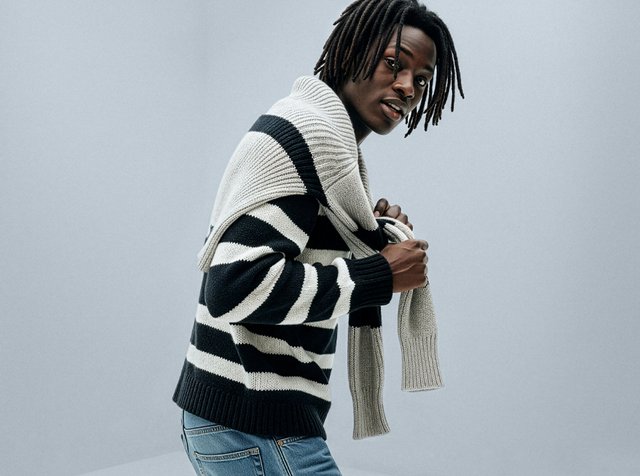 Randig tröja i svart och vitt ull, med ribbad krage och muddar, kombinerad med en ljusgrå halsduk som är draperad över axlarna.