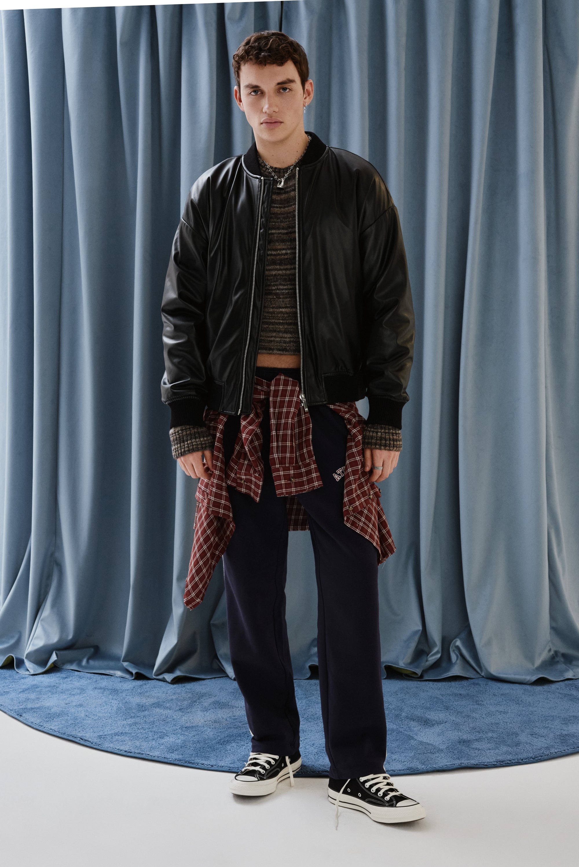 Joven de pie frente a cortinas azules, vistiendo una chaqueta bomber negra, suéter a rayas, camisa de cuadros amarrada a la cintura y pantalones oscuros.