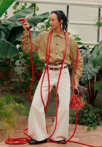 Person in gestreiftem Hemd und weiter Hose hält einen roten Gartenschlauch und eine Vintage-Laterne, umgeben von großen grünen Pflanzen in einem Gewächshaus.