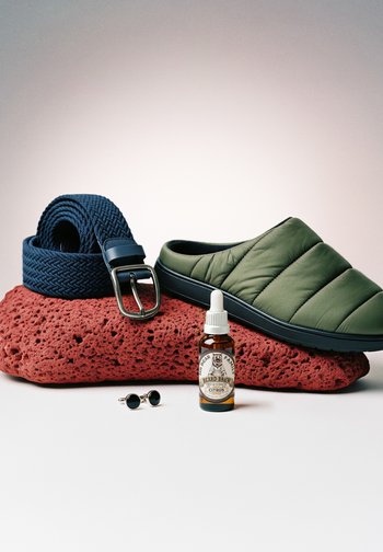 Chaussure à enfiler matelassée verte, ceinture tissée bleue, bouteille d'huile pour barbe et boutons de manchette noirs disposés sur une pierre rouge texturée.