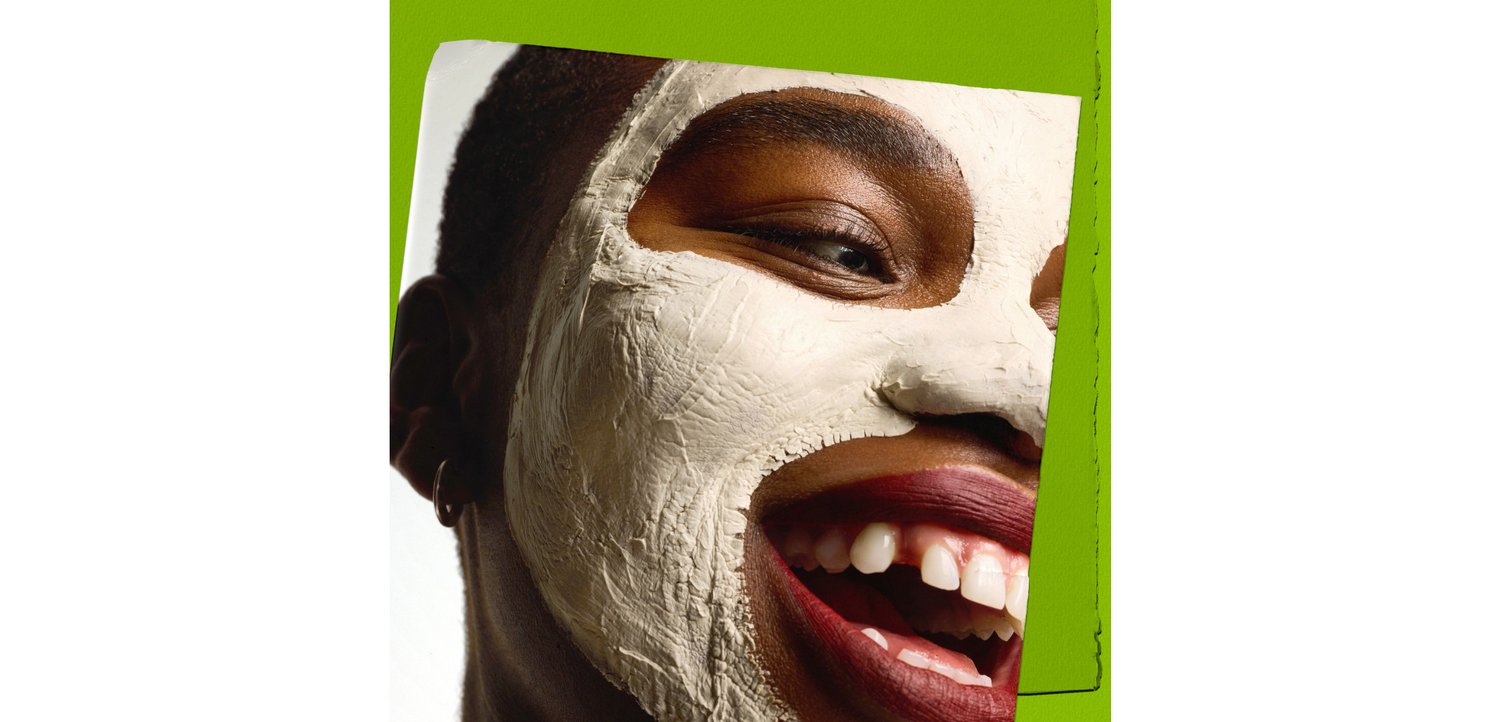 Une personne souriante avec un masque facial en argile blanche sur la peau. L'arrière-plan présente un accent vert vif. Le teint est marron foncé.