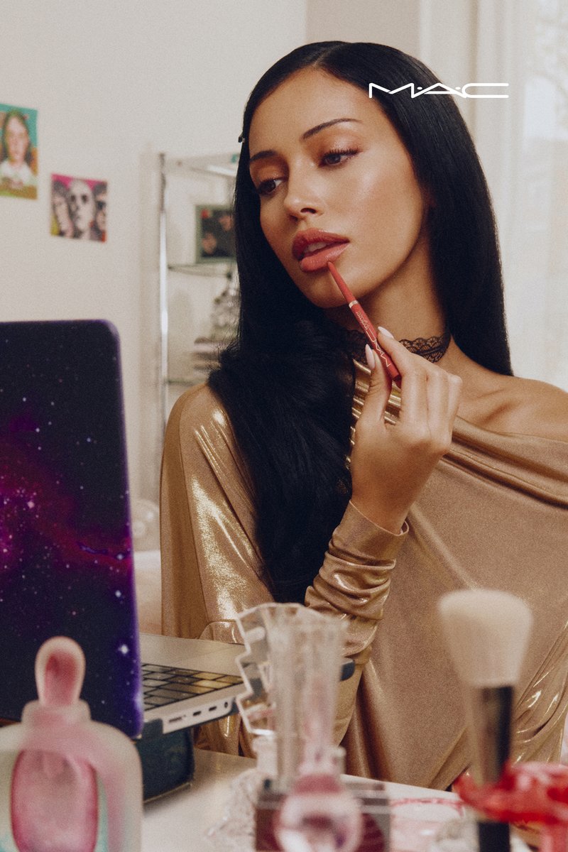 Aplicación de lipstick MAC con una mujer vistiendo un top metálico dorado sin hombros, sosteniendo un delineador de labios rojo. Se ven una laptop y varios productos de maquillaje.
