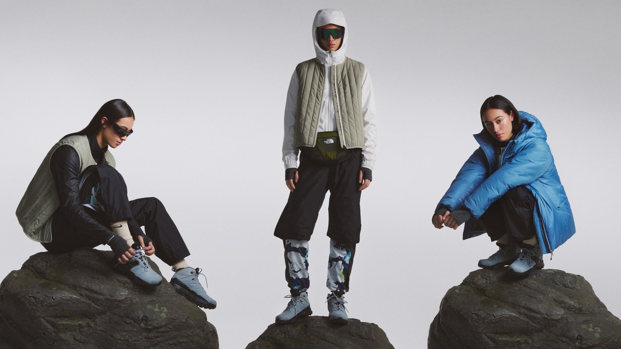 Three models pose on large rocks, showcasing outdoor apparel. One wears a blue puffer jacket, another a beige vest, and the third ties shoes.