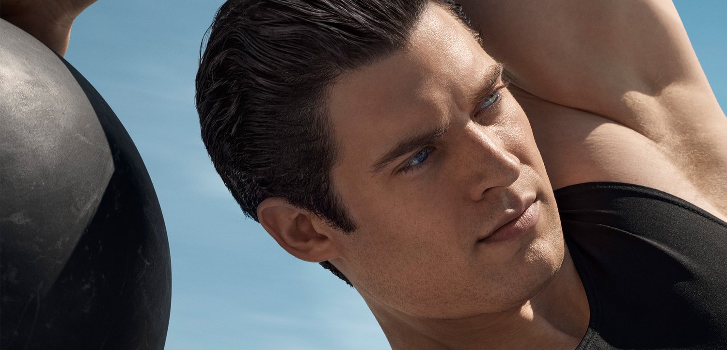 Mandlig atlet med mørkt hår og blå øjne fokuserer på at løfte en tung bold mod en lys blå himmel, hvilket understreger beslutsomhed og styrke.