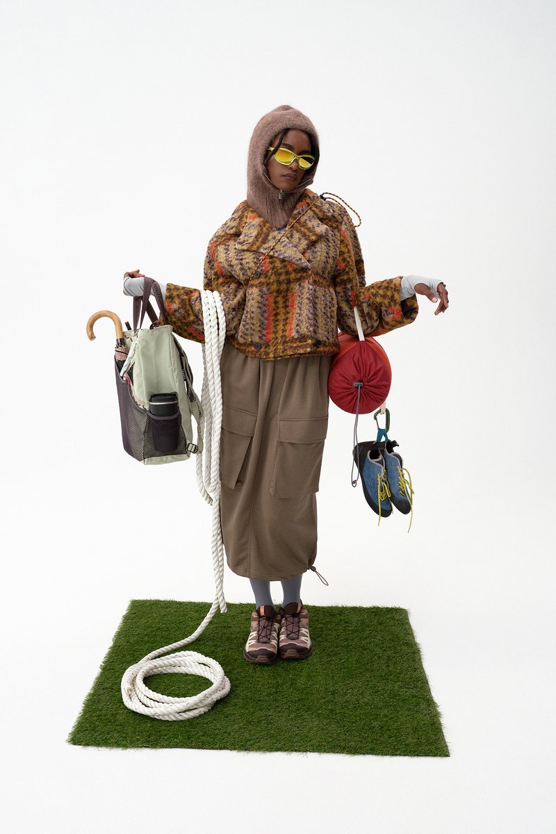 Una persona con piel marrón claro lleva una chaqueta de estampado y gafas de sol amarillas, sostiene bolsas, cuerda y zapatos, de pie sobre hierba verde.