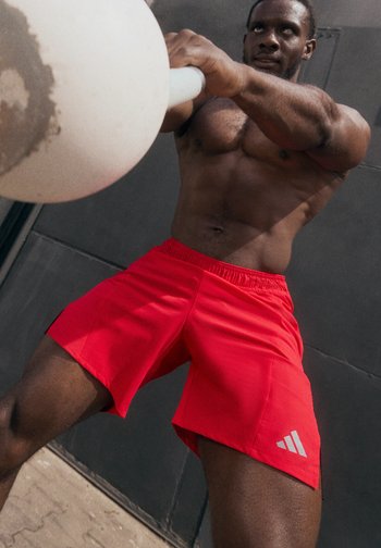 A muscular man in red shorts lifts a large weight overhead, showcasing strength against a dark background.
