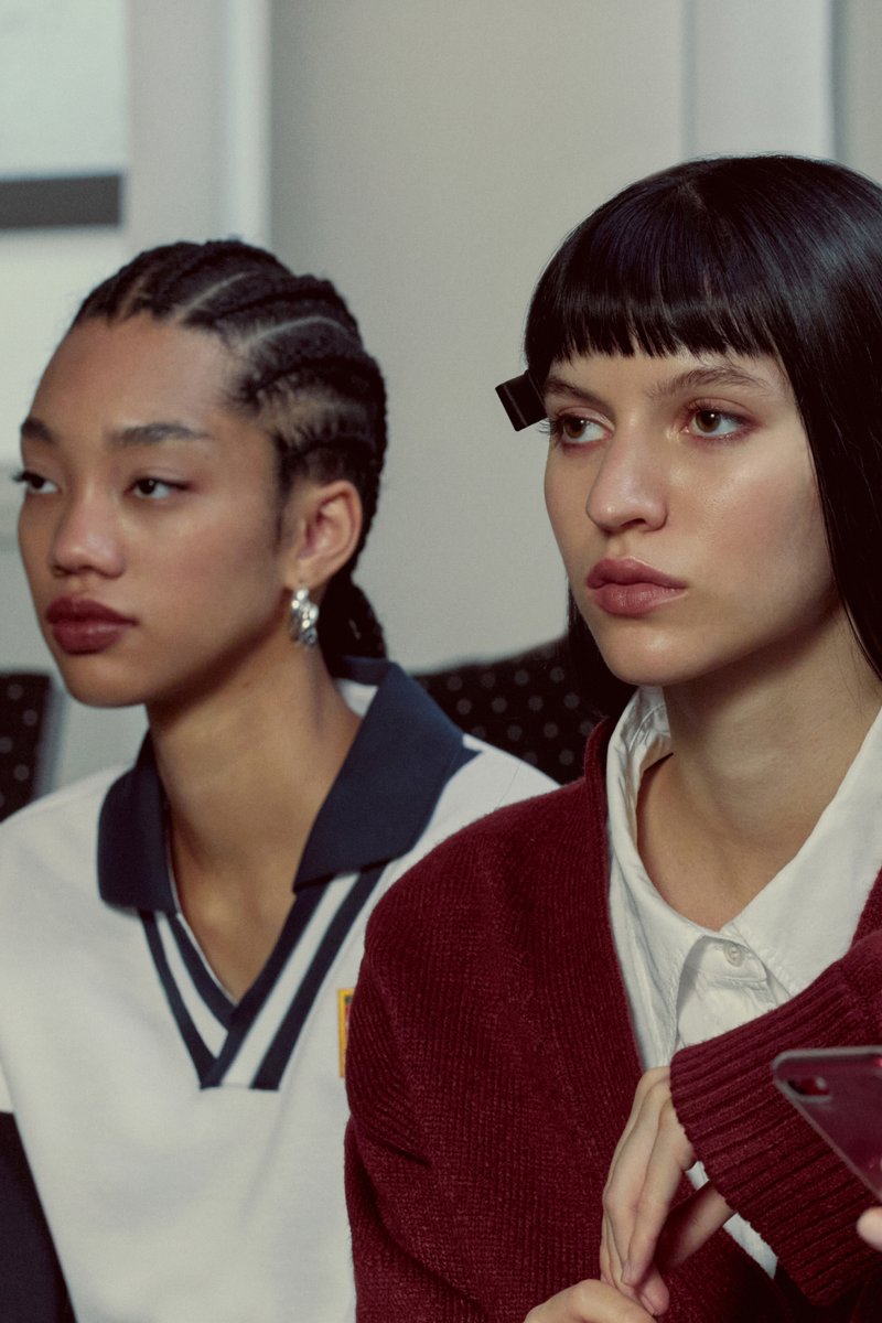 Tre giovani donne siedono affiancate, vestite con abiti casual e alla moda, mentre una tiene un telefono e un'altra indossa delle cuffie.