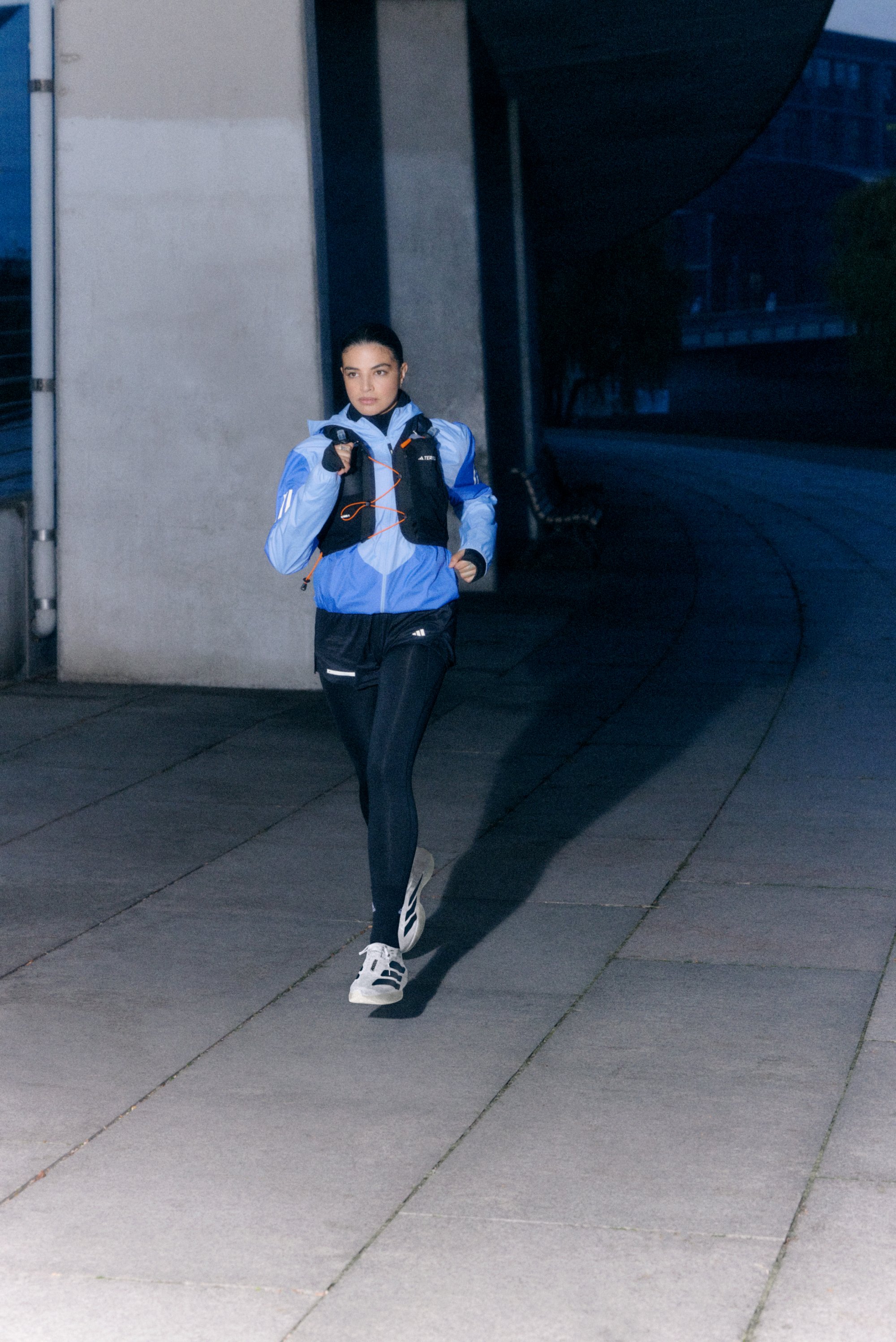 Una mujer corre por un sendero con una chaqueta azul y leggings negros, iluminada por la luz de la tarde, con una sombra proyectada en el suelo.
