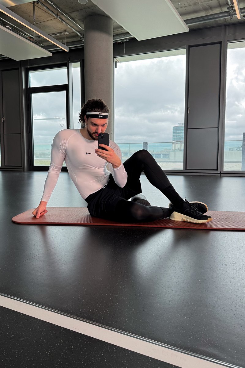 A person in athletic wear sits on an exercise mat, holding a phone and looking at their reflection in a fitness studio with large windows.