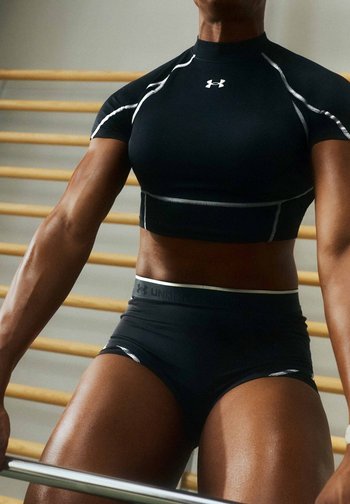 Personne musclée portant une tenue de sport noire Under Armour, tenant une barre d'haltérophilie dans une salle de sport avec des barres murales en bois en arrière-plan.