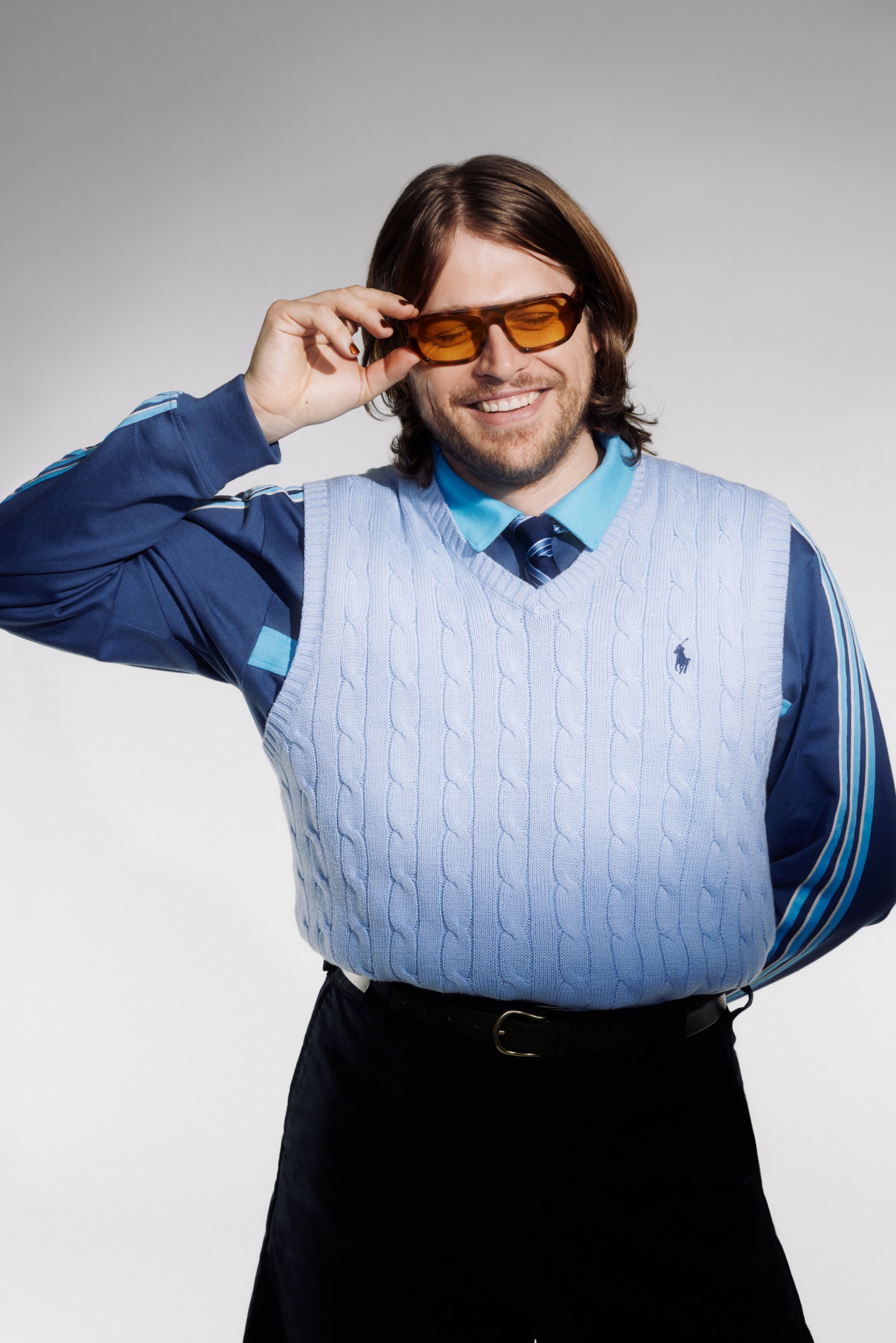 Un hombre con pelo largo sonríe mientras ajusta sus gafas de sol naranjas. Lleva un chaleco de punto azul sobre una camisa azul y una corbata.
