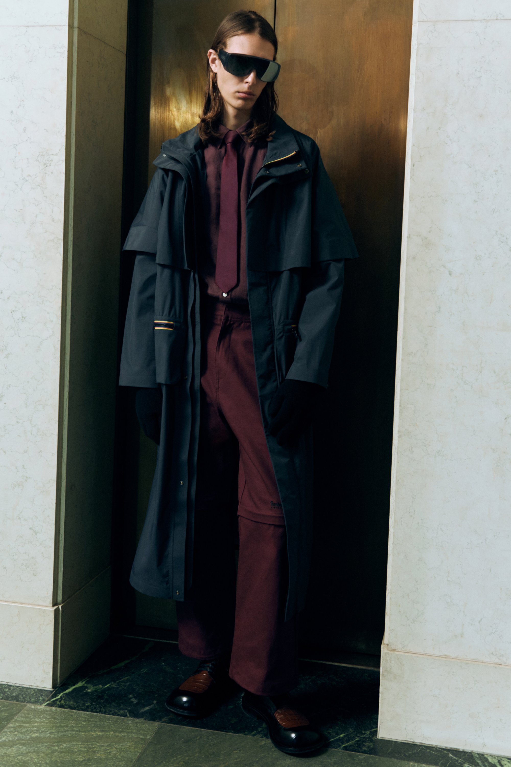 Langer, dunkelblauer Regenmantel mit strukturierten Ärmeln, ein kastanienbraunes Hemd und Hose, schwarze Handschuhe und glänzende braune Schuhe. Schwarze Sonnenbrille vervollständigen den Look.