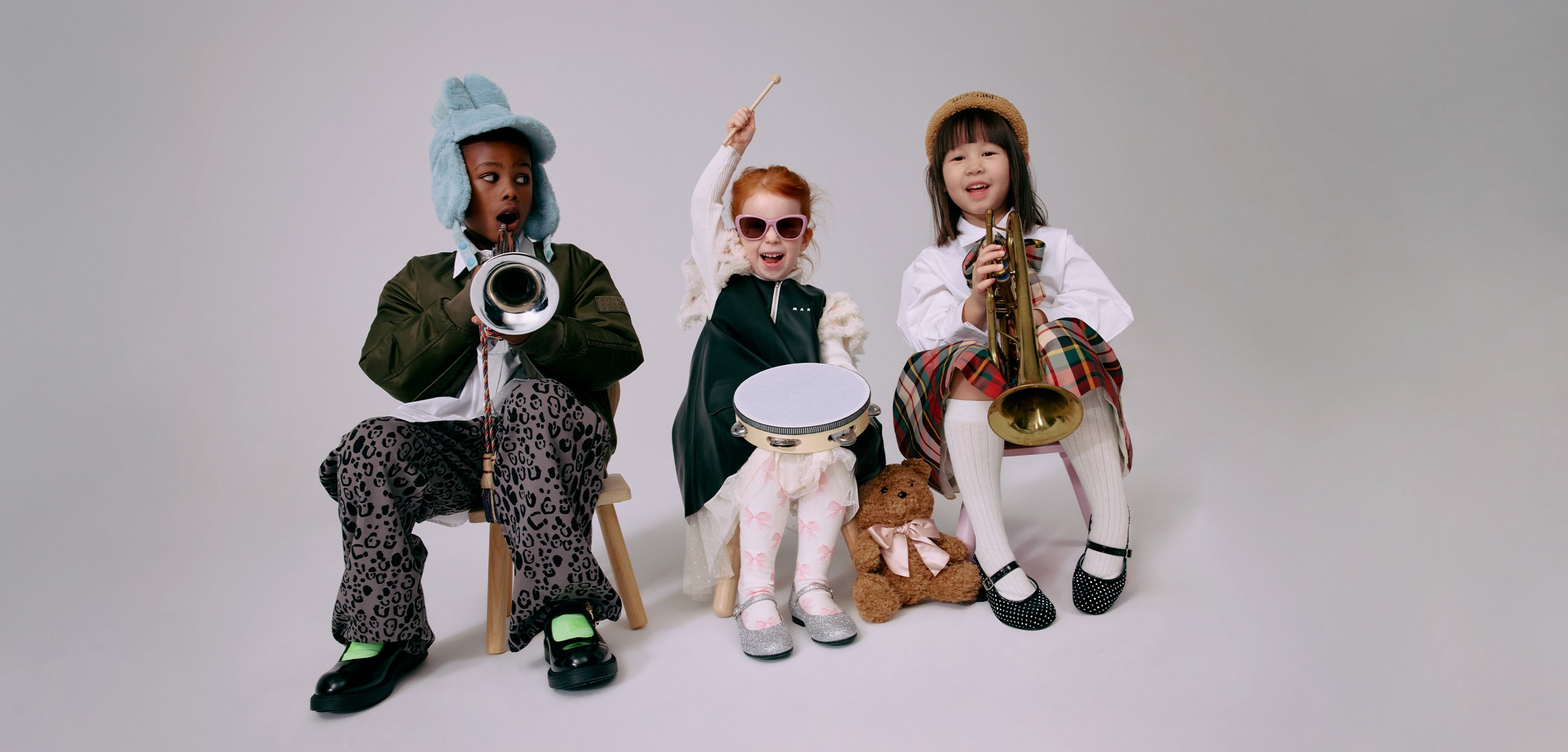 Τρία παιδιά παίζουν μουσικά όργανα: μια τρομπέτ�α, ένα τύμπανο και ένα ταμπουρίνι. Φορούν πολύχρωμες ενδυμασίες, καπέλα και αξεσουάρ με διάφορα σχέδια.