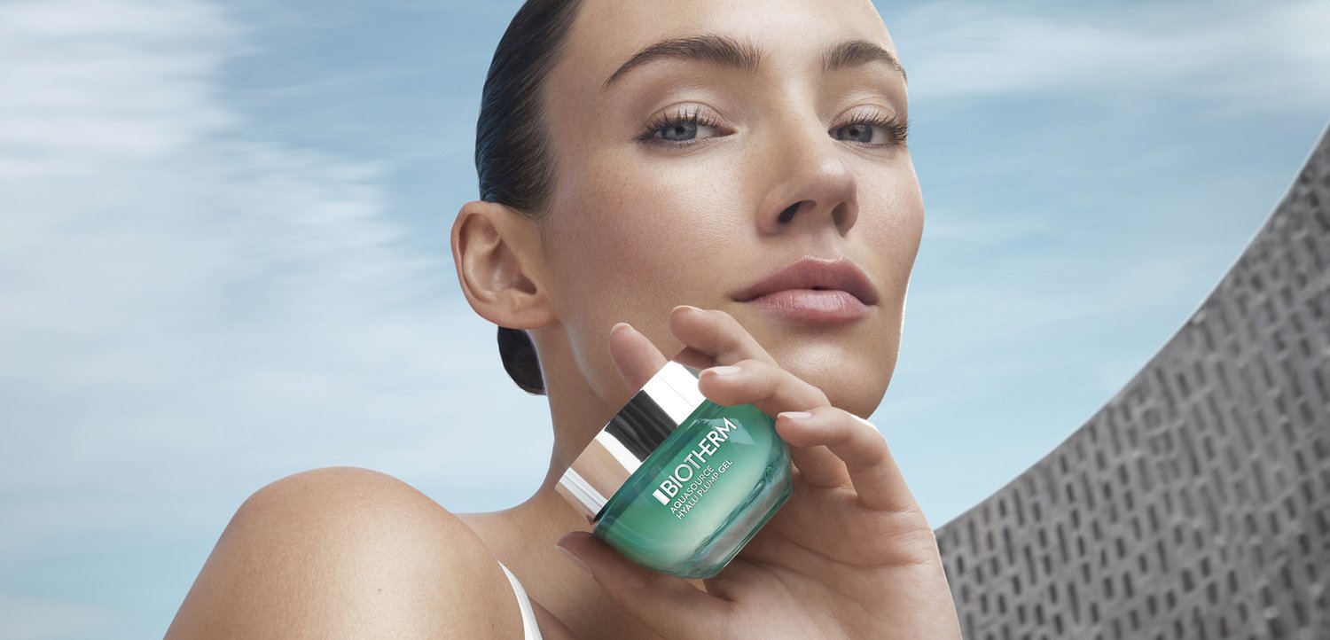 Een vrouw houdt een pot Biotherm Aquasource Hydrating Gel dicht bij haar gezicht, met een moderne architecturale achtergrond en een heldere blauwe lucht.