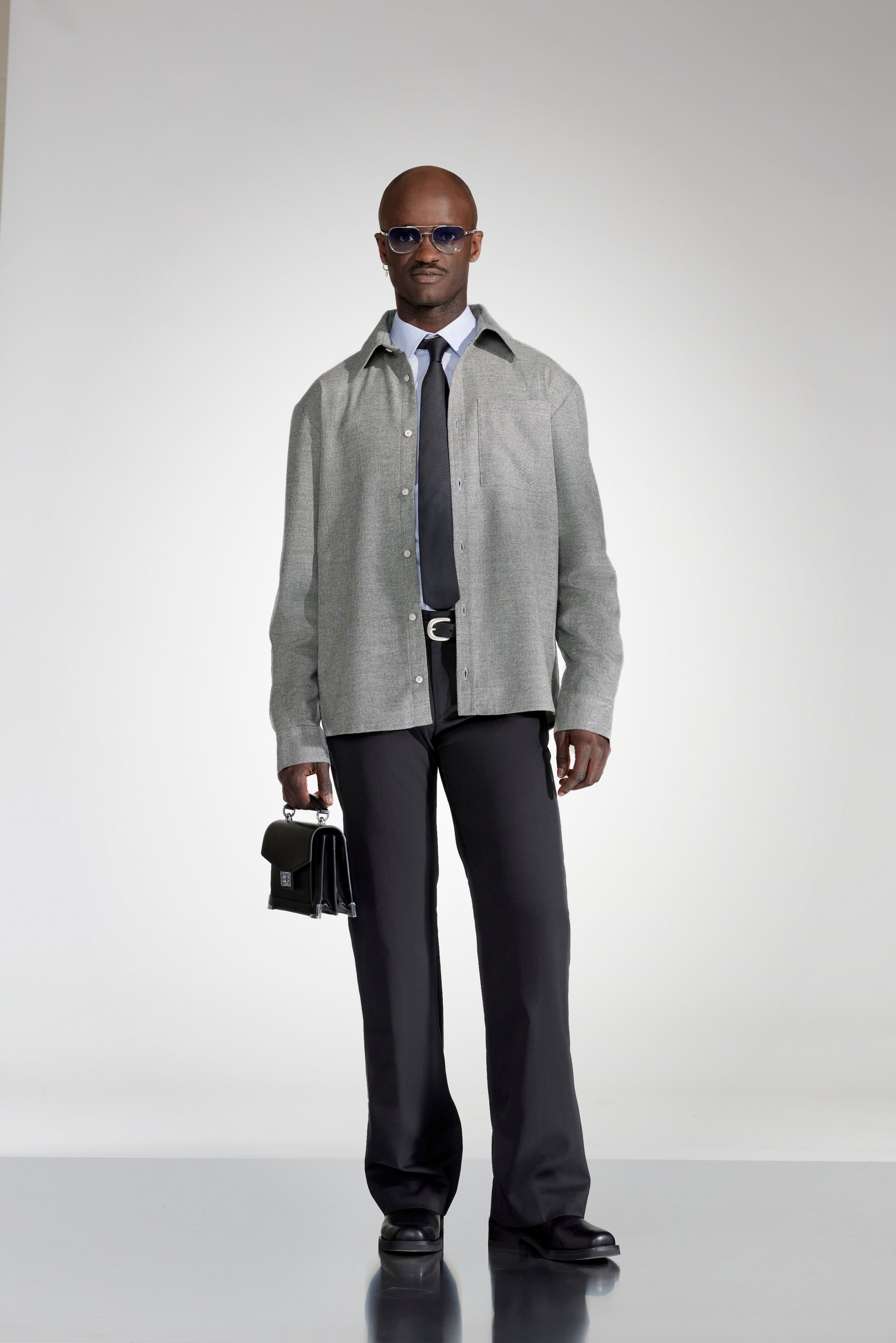 A man stands confidently, dressed in a grey button-up shirt, black tie, and dark trousers. He holds a black briefcase and is wearing sunglasses.