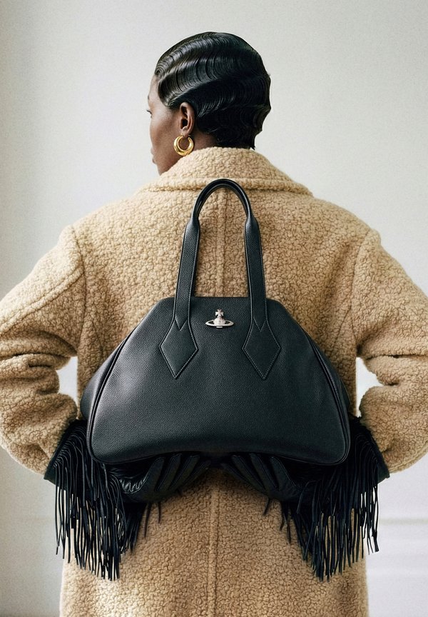 Άτομο με μπεζ παλτό με υφή και κοντά κουρεμένα μαλλιά, φορώντας χρυσούς κρίκους στα αυτιά, κρατώντας μαύρη δερμάτινη τσάντα και μαύρα γάντια με κρόσσια πίσω από την πλάτη.