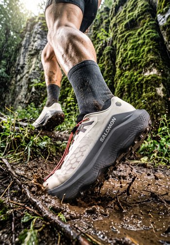Leg muscular legs wearing black socks and white Salomon trail running shoes splash through muddy forest terrain during a wet hike.