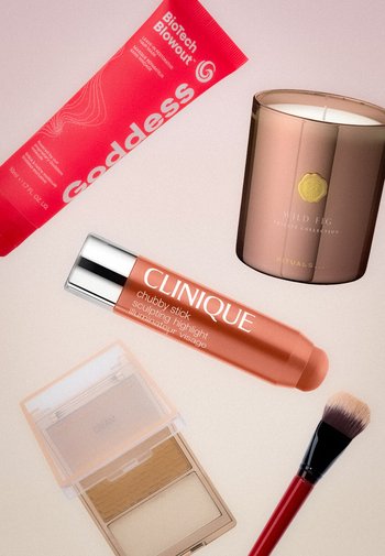 Tube haarmasker Red Goddess, Clinique sculptingshighlight-stick, geurkaars Wild Fig, poederdoosje en make-upkwast op een beige oppervlak.