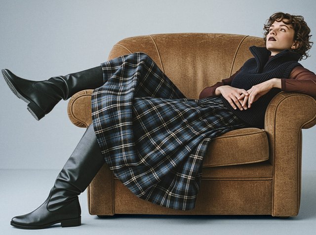 Svarta knähöga stövlar, marinblå stickad väst och rutig kjol med blå, gröna och vita mönster. Modellen lutar sig tillbaka på en brun tygsoffa.