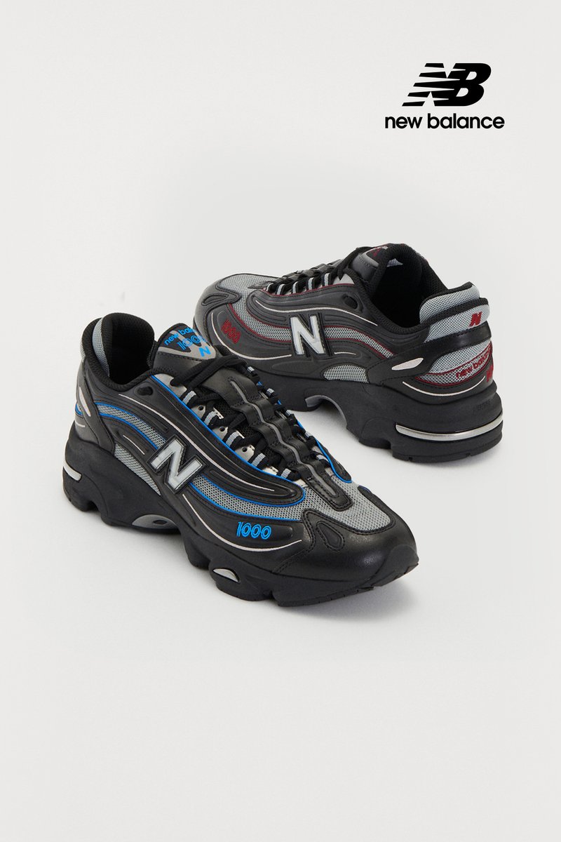 Scarpe da ginnastica New Balance nere e grigie con accenti blu e rossi, caratterizzate da un logo prominente e dalla scritta '1000' sul lato, esibite su uno sfondo bianco.