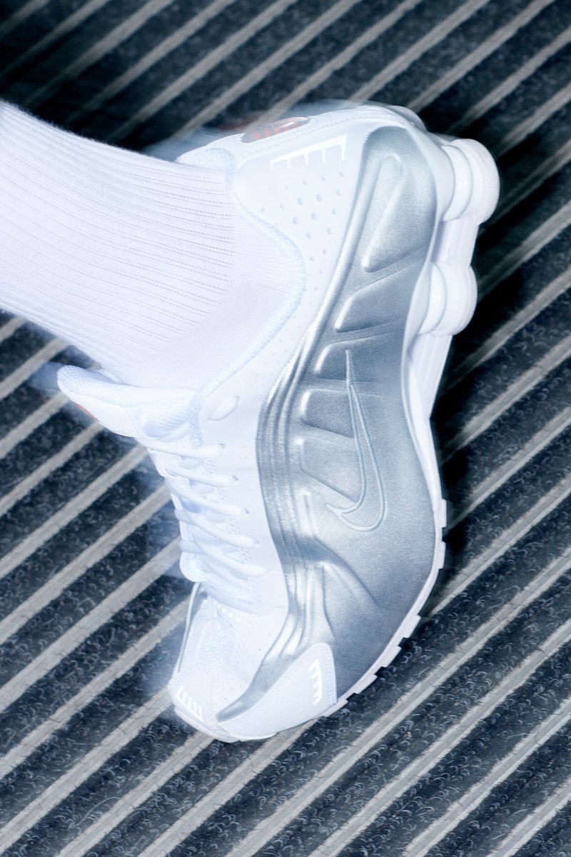 Close-up of a white Nike sneaker with silver accents, resting on a textured black and gray surface. Focus on shoe's details and design.