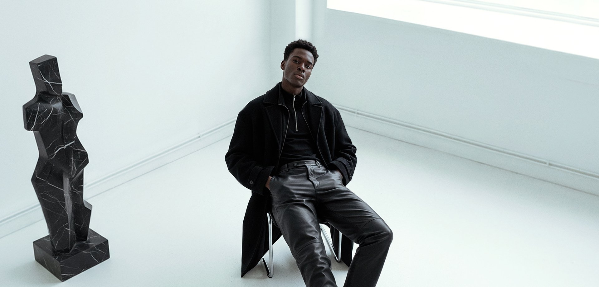 Black leather trousers and a black zip-up top, matched with a long black coat. A geometric black marble sculpture is located nearby on a white surface.