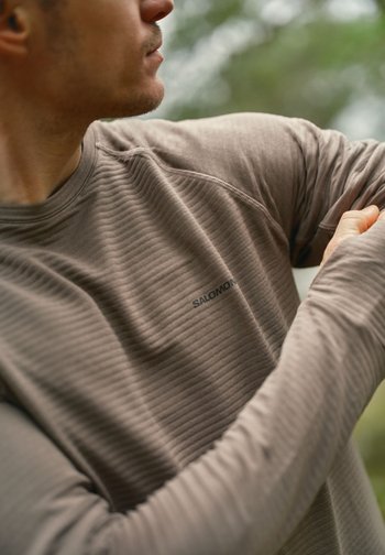 Mann som justerer ermet på en teksturert Salomon langermet skjorte utendørs med uskarpt grønt bakgrunn.