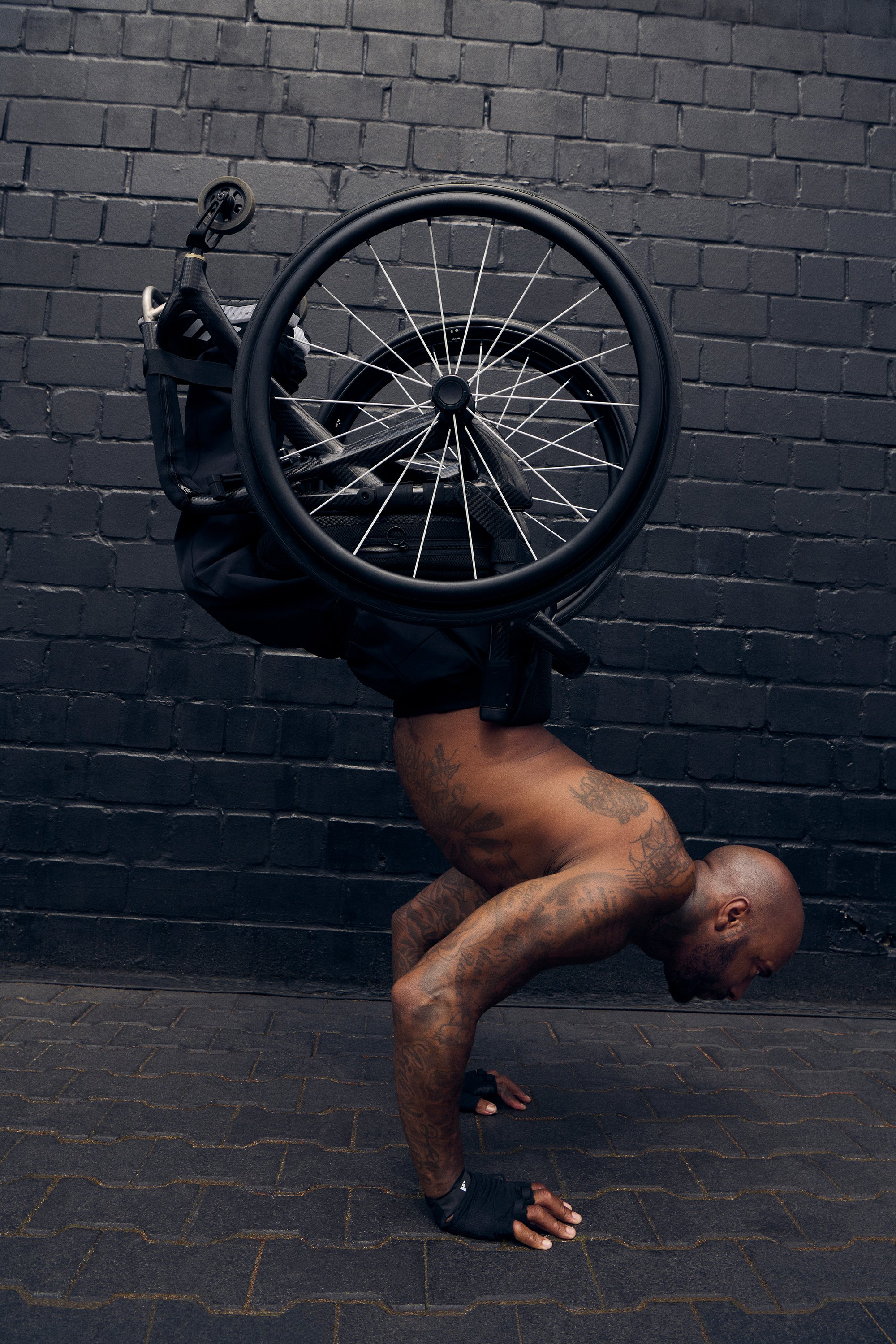 Un hombre musculoso con tatuajes realiza un pino sobre sus manos, equilibrando una silla de ruedas en su espalda, contra una pared oscura y texturizada.