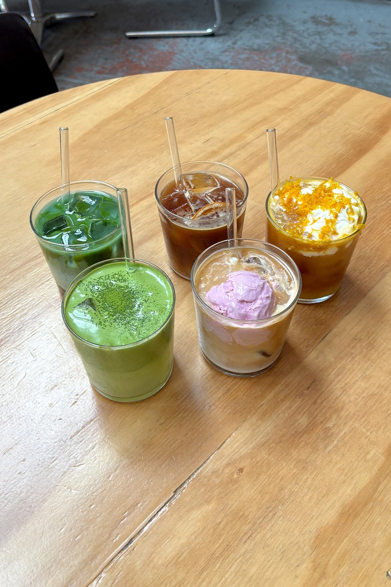 Vijf heldere glazen gevuld met kleurrijke dranken: groene matcha, bruine ijskoffie, oranje drankje met slagroom, roze ijscoupe en een lichtbruine drank.