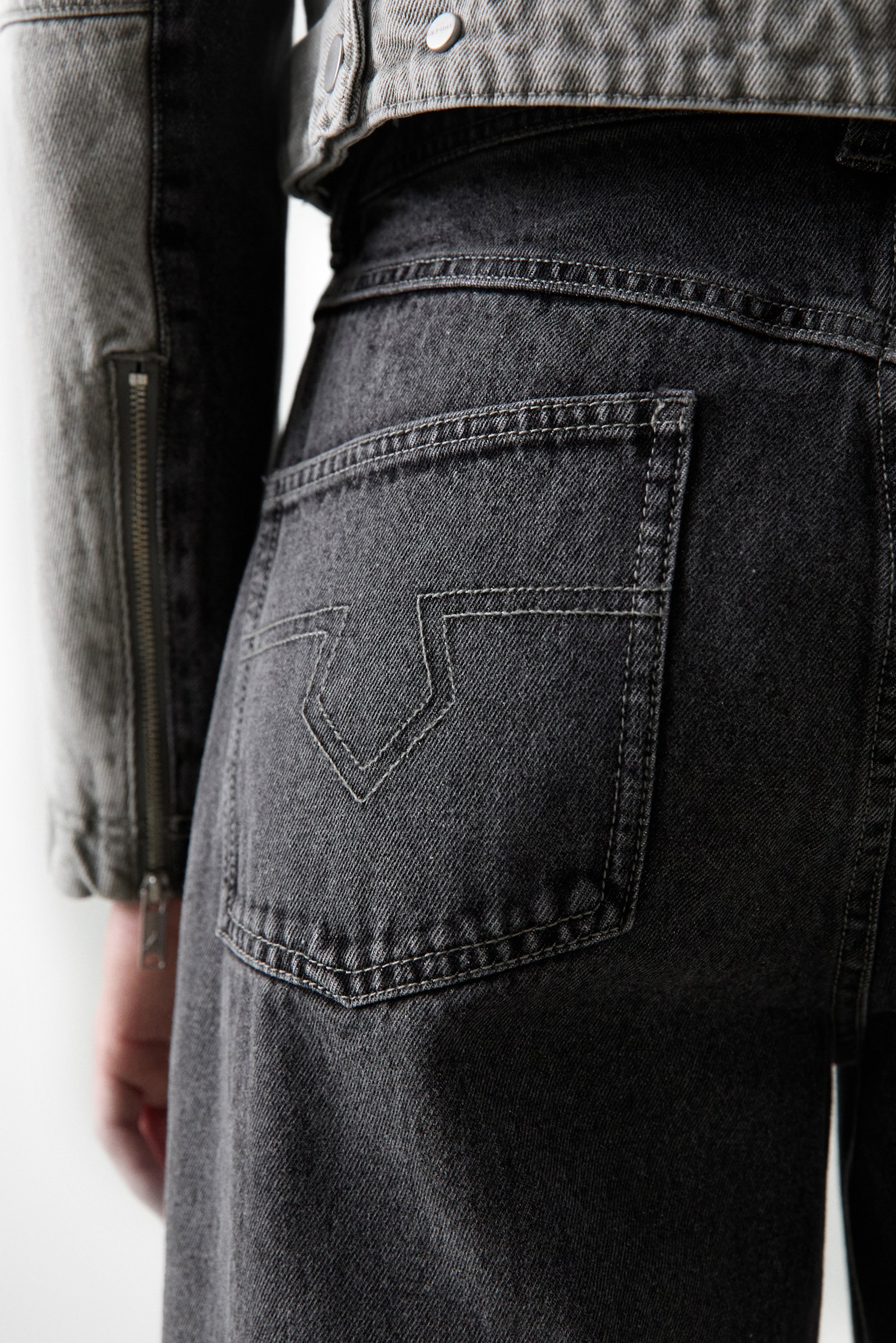 Primo piano della parte bassa della schiena e del fianco di una persona, che indossa jeans grigio scuro con un design di cucitura distintivo sulle tasche, abbinati a una giacca leggera.