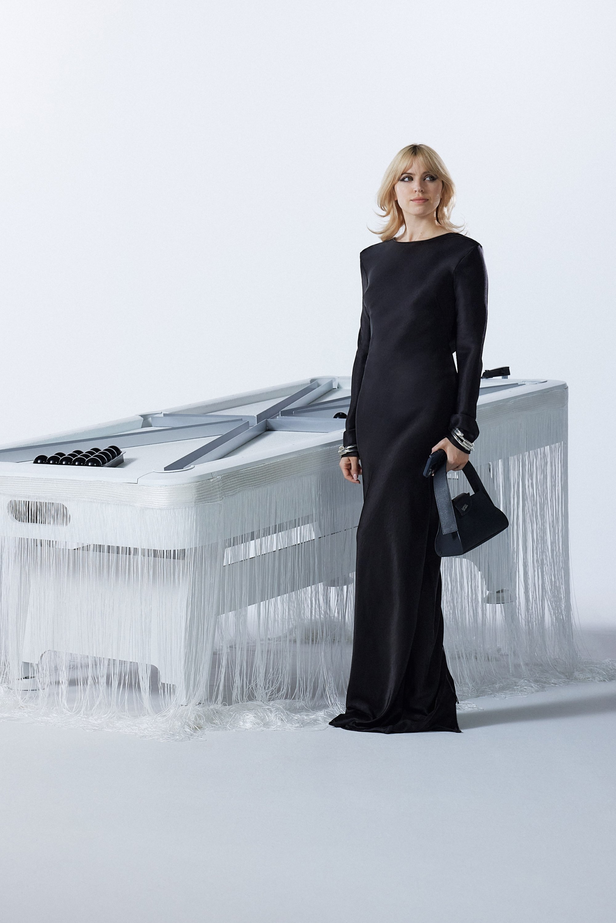 Una mujer con un elegante vestido negro se encuentra junto a una mesa blanca única con flecos, sosteniendo un bolso negro. El fondo es minimalista.