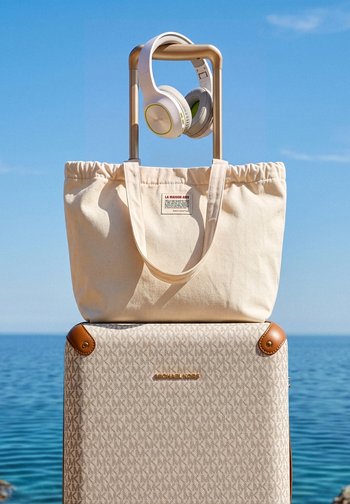 Valise beige Michael Kors avec coins en cuir, sac fourre-tout crème sur la poignée et écouteurs blancs circumauriculaires suspendus à la poignée de la valise, mer en arrière-plan.