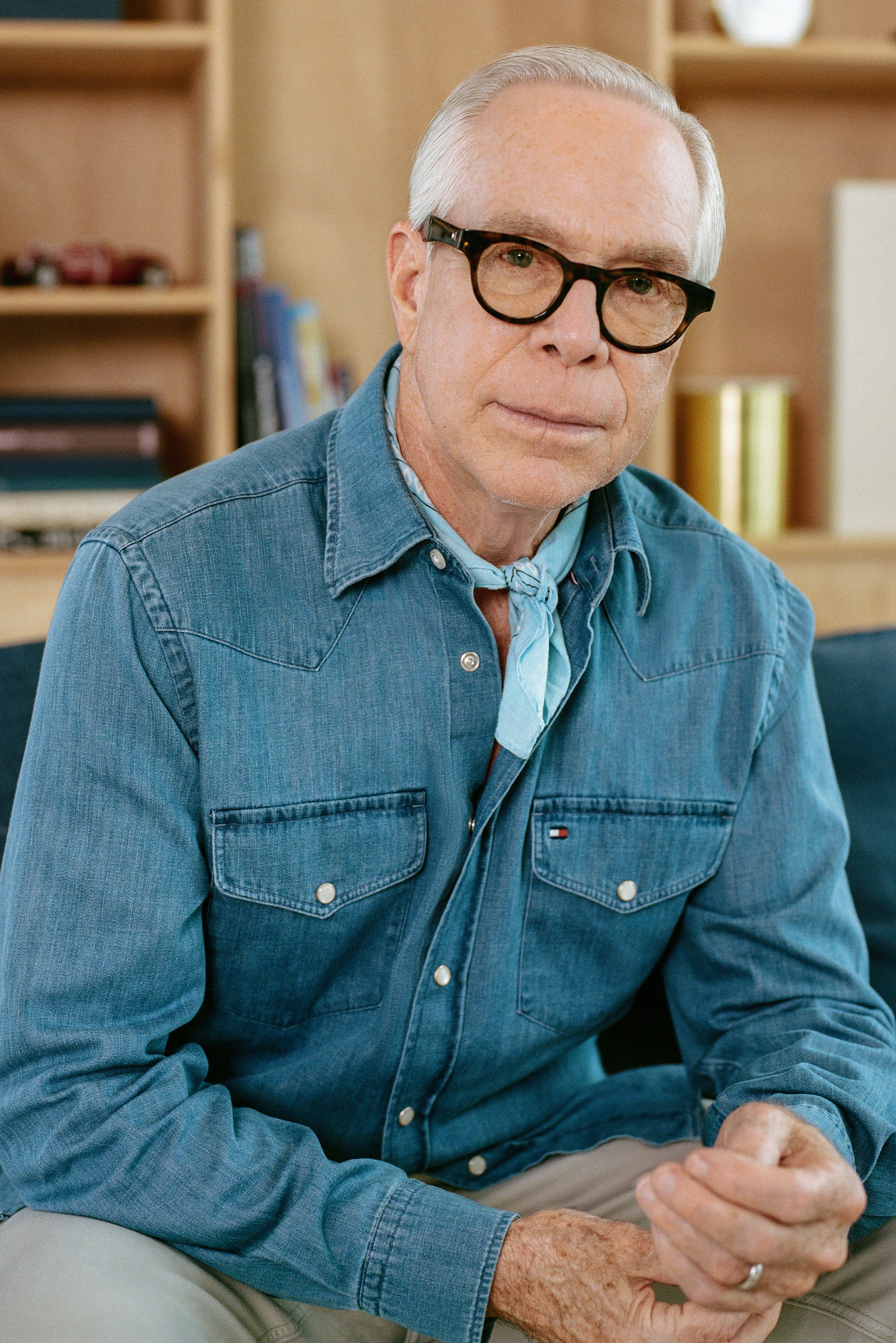 Jeanshemd met lange mouwen, lichtblauwe kleur en twee borstzakken. Draagt een lichtblauwe halsdoek en beige broek. Bril op het gezicht.