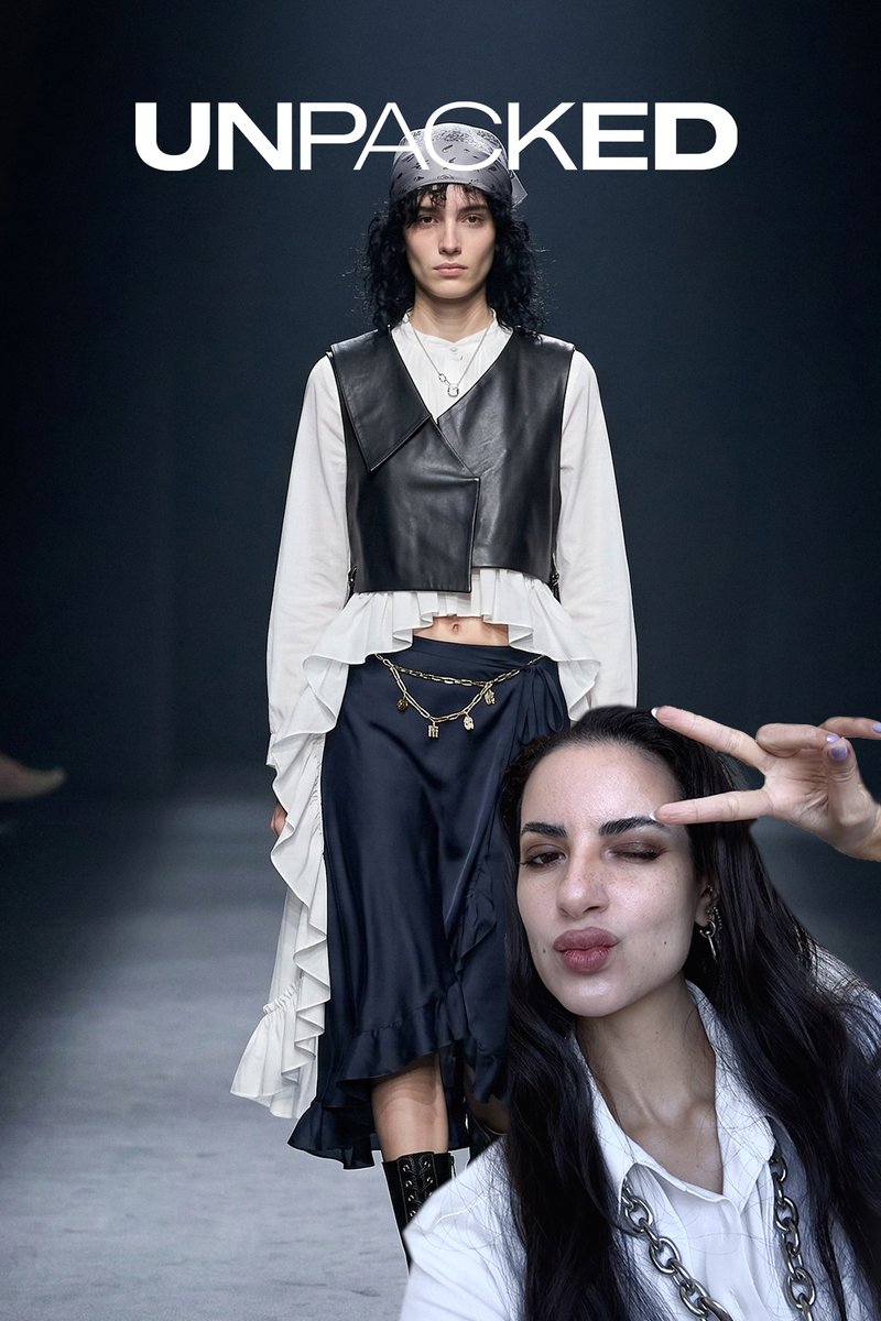 Modelo con blusa blanca con volantes, chaleco de cuero negro, falda azul marino con cadena dorada camina por la pasarela; persona en primer plano hace el signo de la paz.