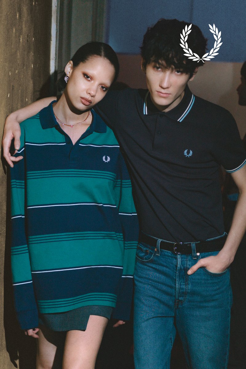 Young man and woman stand side by side; woman wears green striped long-sleeve shirt, man wears black polo and jeans, arm around her shoulder.