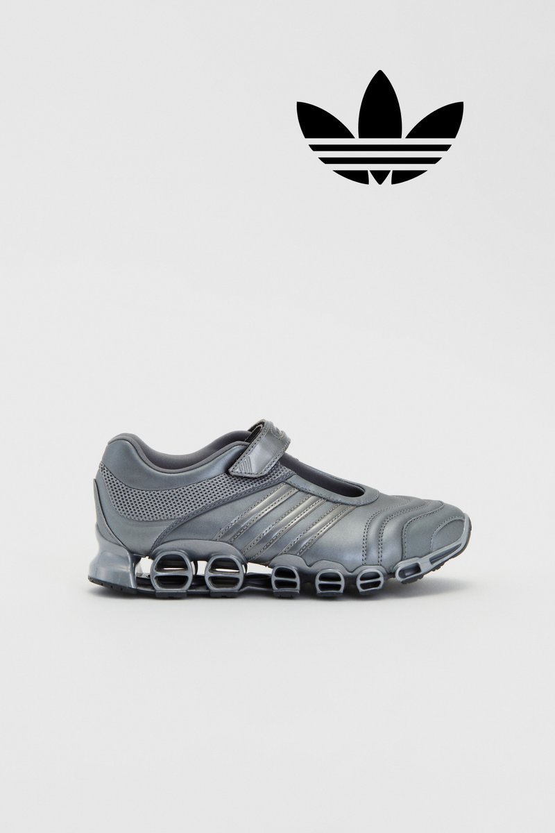 Scarpa sportiva grigia con un design unico e una fascia, caratterizzata da una suola traslucida con elementi di ammortizzazione circolari distintivi. Logo Adidas nero sopra.