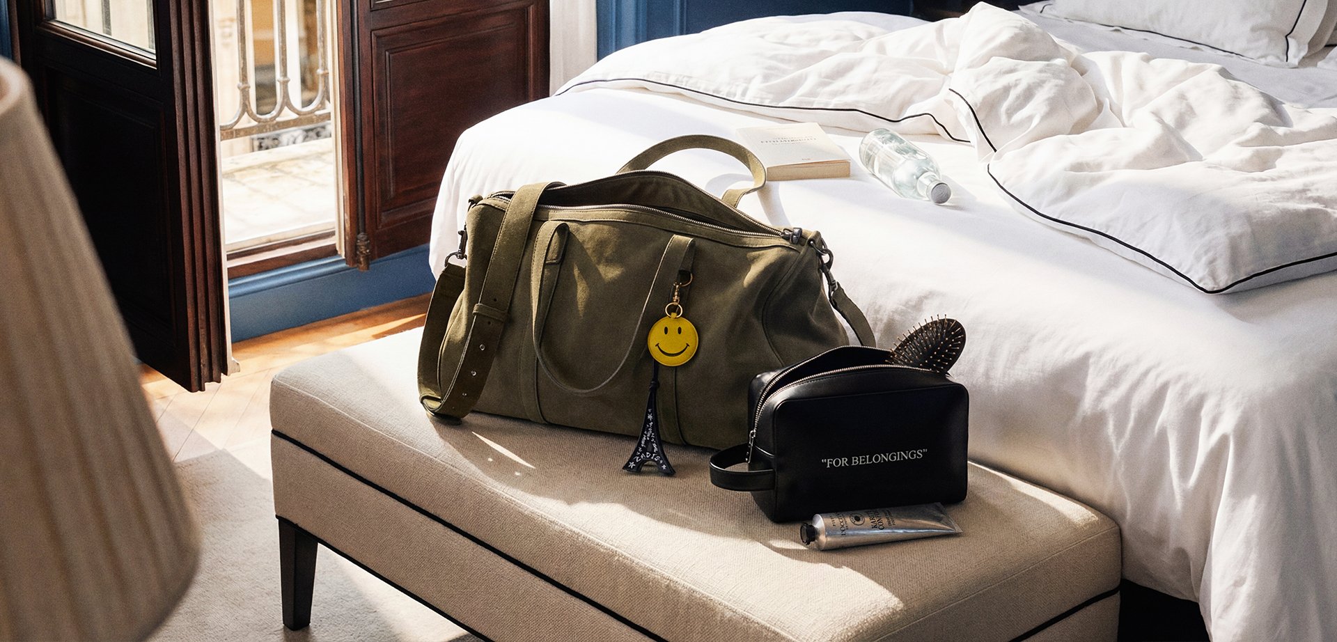 Πράσινη ελιάς τσάντα duffel και μαύρη τσάντα καλλυντικών με μπρελόκ με χαμογελαστό πρόσωπο σε μπεζ πάγκο δίπλα στο κρεβάτι με λευκά σεντόνια και ανοιχτό παράθυρο που αφήνει το φως να μπει.