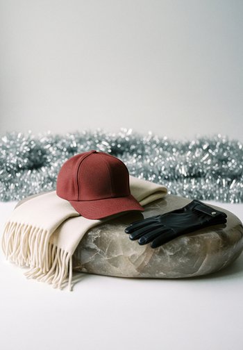 Rdeča kapa z teksturirano tkanino, bež šal z resicami in črne usnjene rokavice na gladki kamenini. Srebrna tinsel v ozadju.