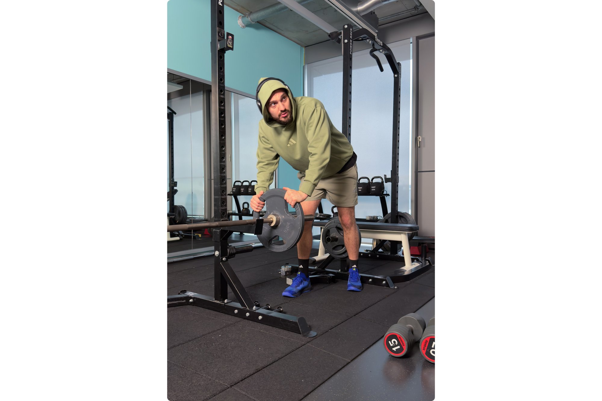 Un hombre con un hoodie verde y pantalones cortos ajusta los pesos en una barra en un gimnasio, con mancuernas visibles en el suelo.