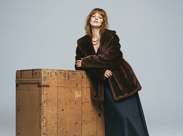 Brun pälsjacka i konstmaterial med bred krage, ihop med en klänning i marinblått. En träback med synliga märken är i förgrunden.