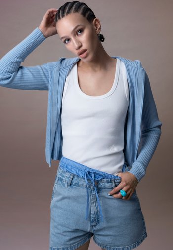 Jeune personne avec des tresses cornrow portant un débardeur blanc, un cardigan bleu clair, un short en jean et une bague turquoise, posant sur un fond neutre.