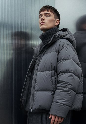 Grå vattert jakke med glidelåslukking og tilhørande hette; har vattert polstring og høy krage. Strukturert stoff, minimalistisk design.